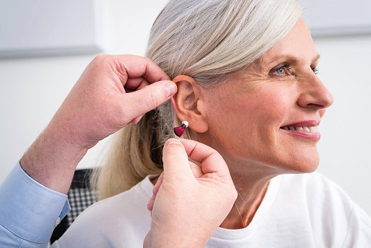 Seniorin bekommt ein modernes Hörgerät angepasst, Hörakustiker hilft beim Einsetzen.